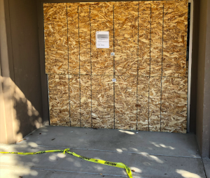 Bank entrance boarded up, with a sign tha says "branch temporarily closed". Yellow Caution tape lies tattered on the ground in front.